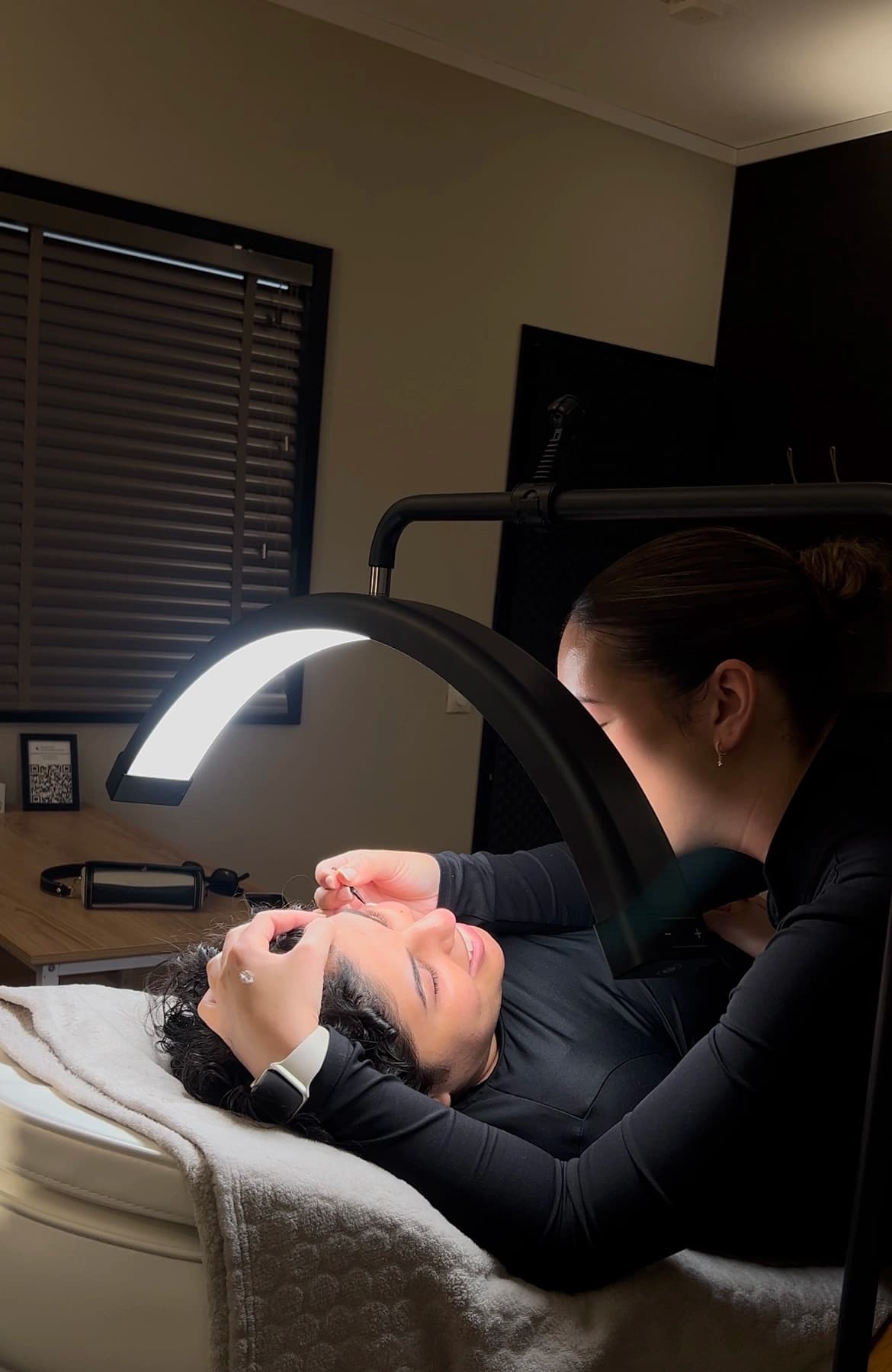 Sarah werkt onder de ringlamp aan de wenkbrauwen van een klant in de salon